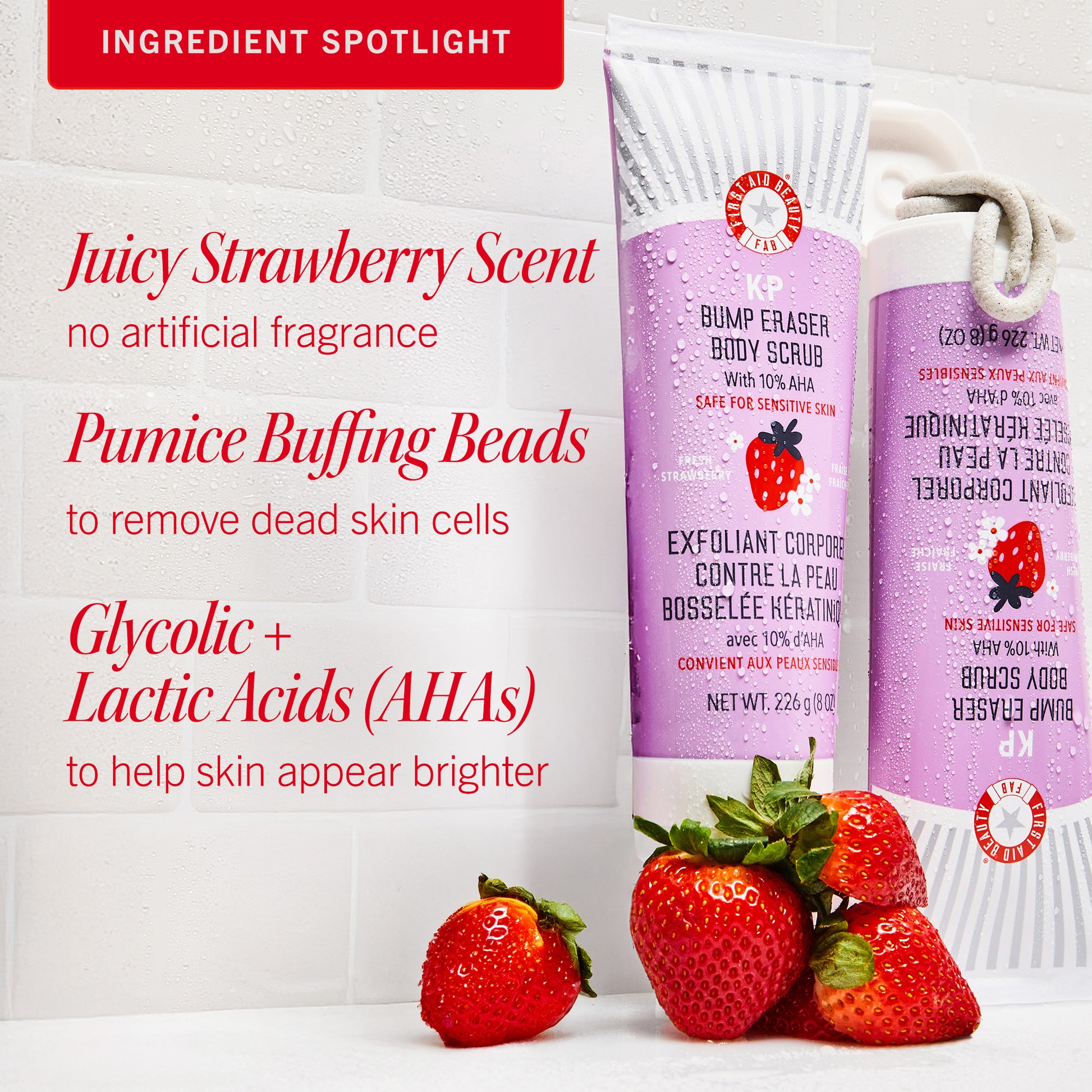 Purple body scrub tubes stand upright, surrounded by strawberries on a wet tiled surface. Text reads: "Juicy Strawberry Scent, Pumice Buffing Beads, Glycolic + Lactic Acids (AHAs)."