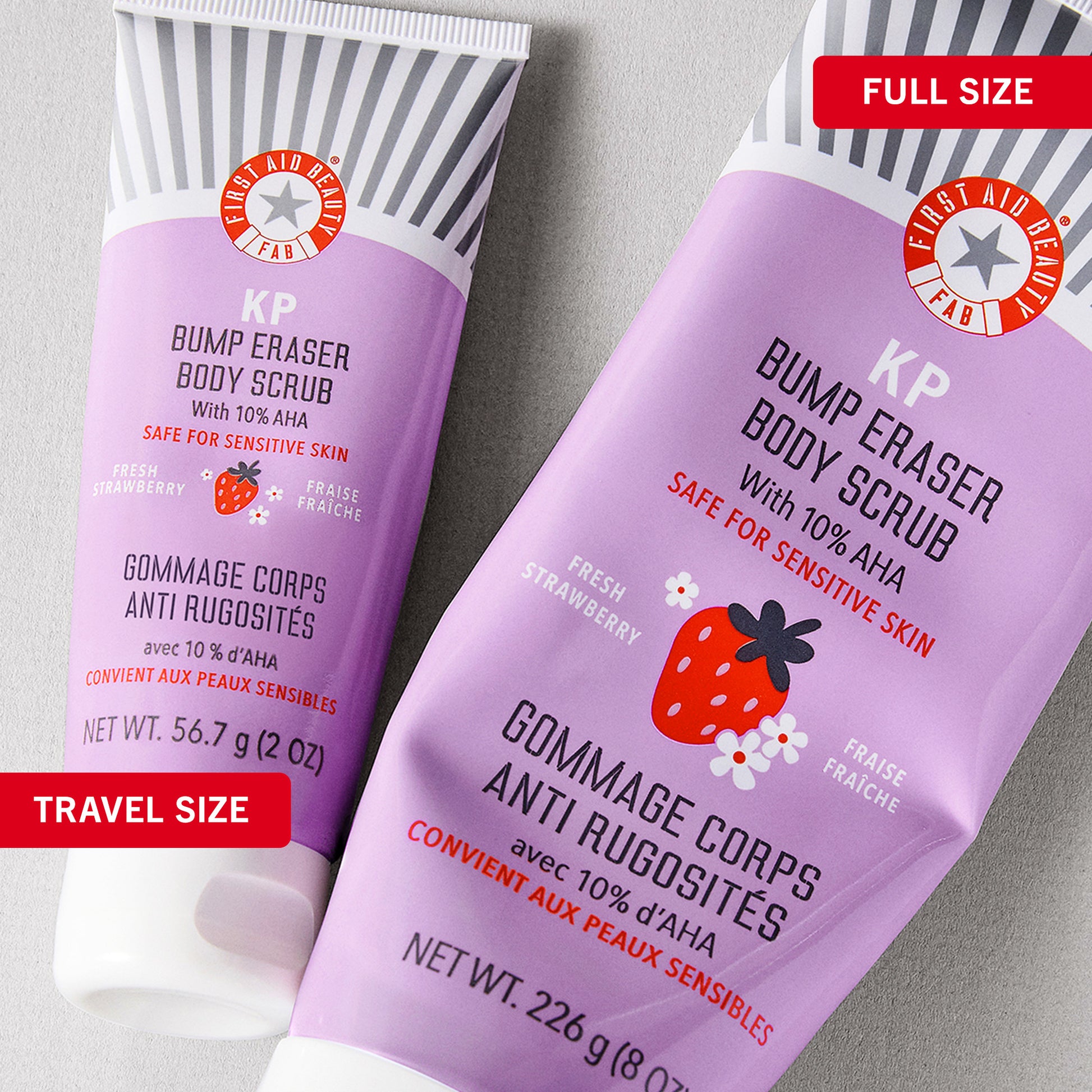 Two purple tubes labeled as "First Aid Beauty KP Bump Eraser Body Scrub" with 10% AHA and fresh strawberry scent. One is marked "FULL SIZE," the other "TRAVEL SIZE." Featured on a light gray surface.