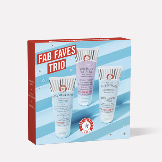 The image shows a skincare gift box labeled "FAB FAVES TRIO" featuring three products: "Ultra Repair Cream," "Bump Eraser Body Scrub," and "Face Cleanser" by First Aid Beauty, with a blue striped background.